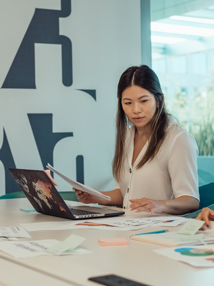 Asian woman in a modern office environment, reviewing paperwork with a laptop on the desk.